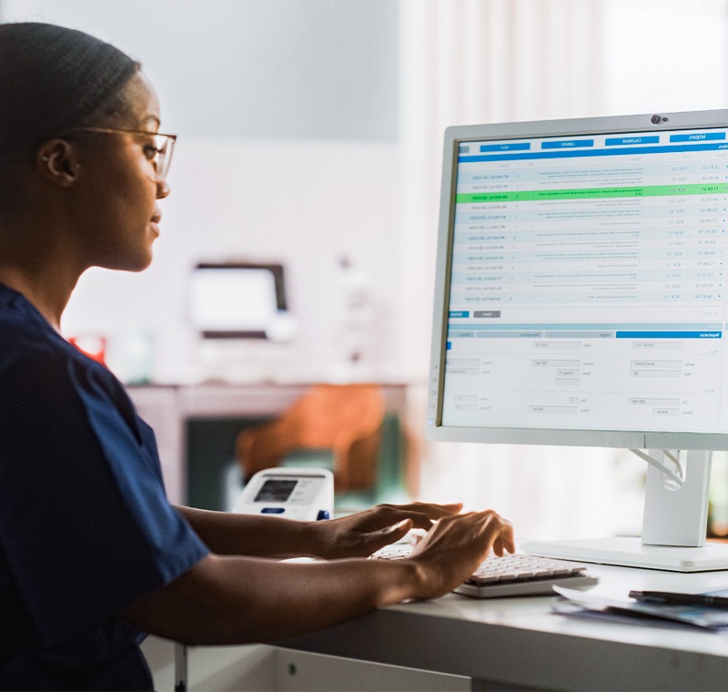 An electronic health records and reimbursement specialist works at a computer in a medical office, reviewing patient billing information and updating digital health records accurately.