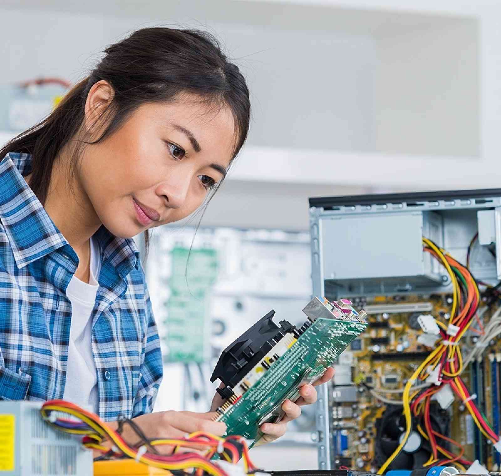 A PC technician works at a computer repair bench, examining internal hardware components of a desktop tower while referencing diagnostic software on a monitor in a modern tech workspace.