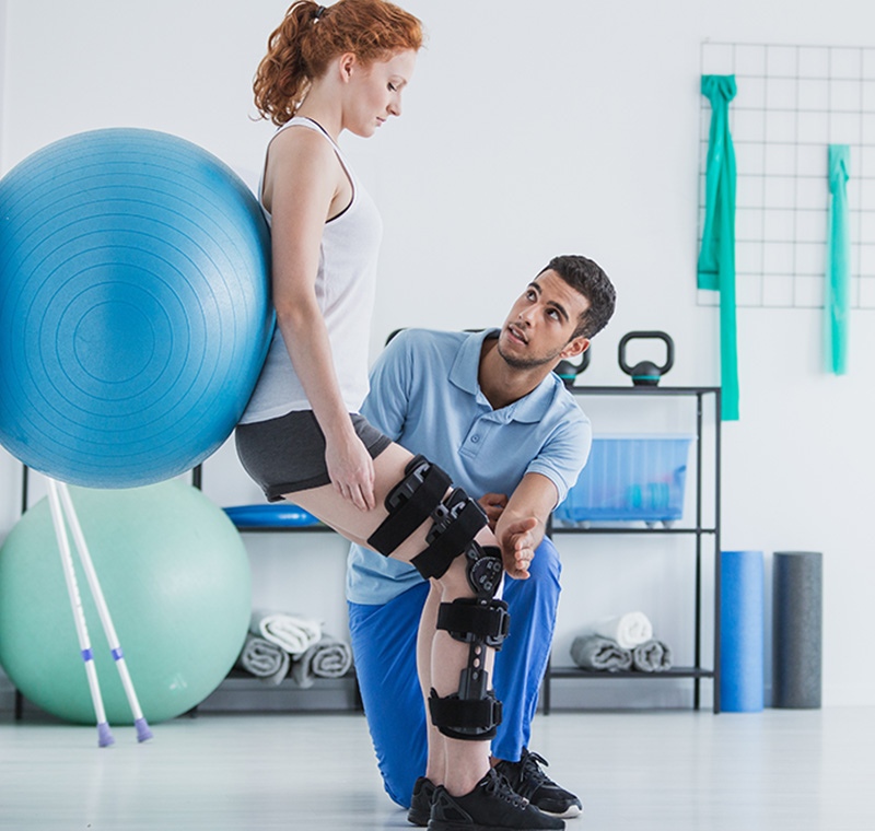 A physical therapy aide and administrative specialist assists a patient with rehabilitation exercises while organizing therapy records in a bright clinical setting.